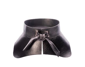 May include: A wide black leather corset belt featuring a textured surface, a structured high-waisted design, and a front tie closure with metallic hardware accents. The belt has a smooth interior lining and a flared shape that tapers at the waist.