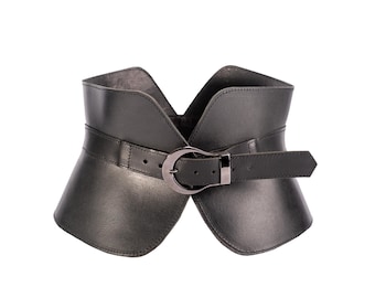 May include: A wide black corset-style belt featuring a smooth finish and a structured, flared silhouette. The belt is secured at the center with a dark metallic buckle and a matching prong, with a pointed end strap that feeds through the buckle loop.