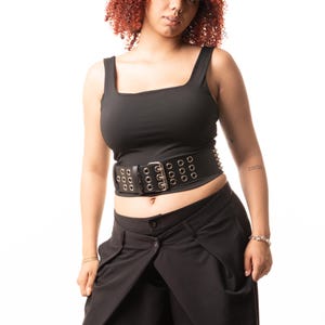 May include: A black sleeveless tank top with a square neckline paired with a wide black belt featuring three rows of silver-toned metal grommets and a central silver buckle, worn over black trousers with a layered, overlapping front panel design.