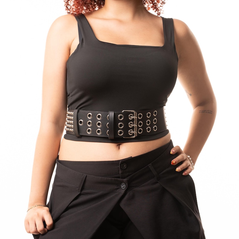 May include: A wide black belt featuring three horizontal rows of silver-toned metal grommets and a matching silver-toned metal buckle. The belt is worn over a black sleeveless top and black trousers, showcasing a structured design with metallic hardware.