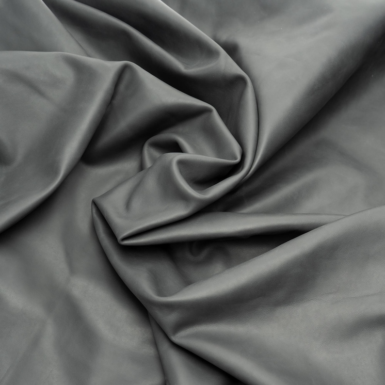 Puede incluir: Una pieza de cuero de color gris oscuro s&oacute;lido con un acabado liso y mate. El material est&aacute; drapeado en pliegues suaves y fluidos, creando sombras y reflejos profundos en la superficie que enfatizan la textura y flexibilidad de la piel.
