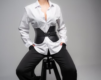 Könnte beinhalten: Ein weißes, langärmeliges Hemd mit Knopfleiste, kombiniert mit einem breiten, schwarzen Korsettgürtel mit mittiger Schnalle. Das Outfit wird mit einer schwarzen Hose getragen, was einen kontrastreichen Look mit klaren Linien und einer strukturierten Silhouette erzeugt.
