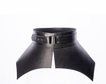 May include: A wide black leather corset belt featuring a structured, flared silhouette. The belt includes a horizontal strap across the front secured with a dark metal rectangular buckle and multiple adjustment holes, creating a defined waist shape.