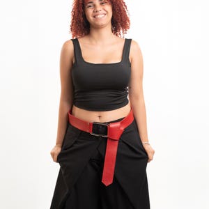 Puede incluir: Un cinturón rojo brillante con una hebilla rectangular de tono plateado y un extremo largo y puntiagudo que cuelga. El cinturón se lleva sobre pantalones negros y se combina con una camiseta negra sin mangas con escote cuadrado y tirantes anchos.