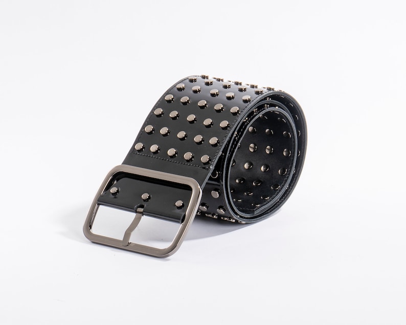 May include: A wide black belt with a glossy finish, featuring multiple rows of silver-toned metal studs across the surface. The belt includes a large, rectangular silver-toned metal buckle and is coiled to show the interior and exterior details.