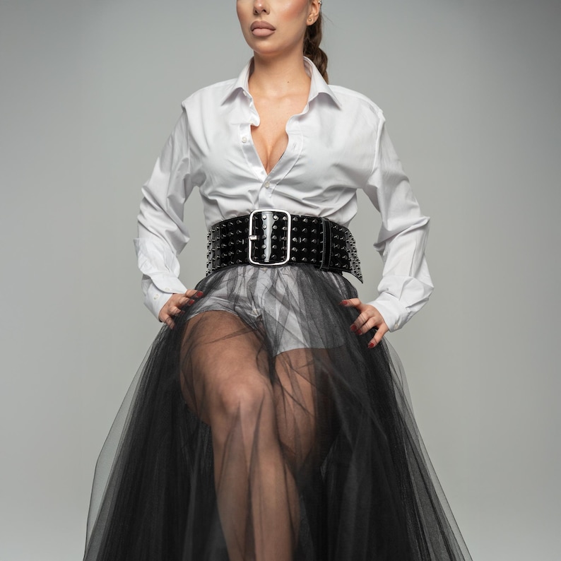 May include: A crisp white long-sleeved button-down shirt paired with a wide black studded belt featuring a large silver buckle. The outfit includes a sheer black tulle skirt layered over white shorts, creating a layered look with contrasting textures.