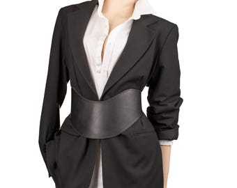 May include: A black blazer worn over a white button-down shirt, cinched at the waist with a wide black belt. The blazer features long sleeves and a notched lapel, while the white shirt has a visible collar and a front button placket.
