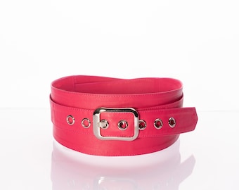 May include: A wide hot pink belt featuring a large, rectangular silver-toned metal buckle and a series of evenly spaced silver-toned metal grommets along the strap. The belt is coiled into a circular shape, showcasing its smooth, solid-colored surface.
