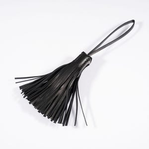 Könnte beinhalten: Eine schwarze Lederquaste mit einer Reihe dünner, vertikaler Streifen, die oben von einer genähten Lederkappe zusammengehalten werden. Eine lange, schmale Schlaufe erstreckt sich von der Oberseite der Kappe und dient als funktionaler Befestigungspunkt.