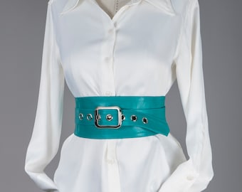May include: A white long-sleeved button-down shirt paired with a wide teal belt featuring a large rectangular silver buckle and silver metal eyelets along the strap. The belt is cinched around the waist over the shirt, which is tucked into blue jeans.