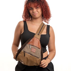 May include: A triangular sling backpack featuring a mix of brown, tan, and olive green panels. The bag includes two front zippered pockets, a single shoulder strap with a metal ring attachment, and a small rectangular leather patch on the front.