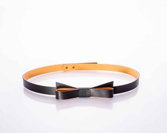 May include: A black belt featuring a decorative bow detail on the front. The belt has a smooth black exterior and a contrasting tan interior lining. It includes a simple adjustment mechanism with visible metal studs for securing the strap length.