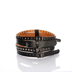 Może przedstawiać: Czarny skórzany pasek z wieloma warstwami i srebrnymi ćwiekami. Pasek ma trzy klamry.