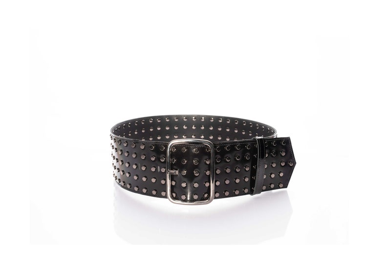 May include: A wide black belt featuring a glossy finish and multiple rows of small, dark metallic studs. The belt is secured with a rectangular silver-toned metal buckle and includes a matching black belt loop to hold the end of the strap in place.