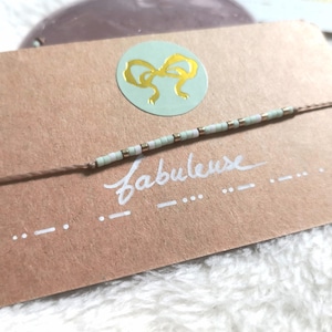 Peut inclure: Une carte marron clair avec un autocollant rond vert clair représentant un nœud doré. Une fine ficelle avec de petites perles blanches et dorées est attachée autour de la carte. La carte dit "Fabuleuse" en écriture cursive blanche.