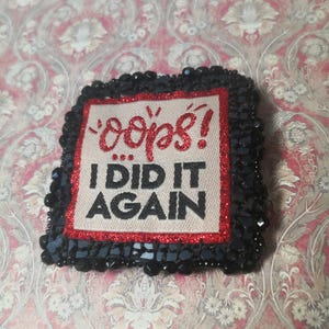 Peut inclure: Un écusson carré avec les mots "oops! I DID IT AGAIN" en rouge et noir. L'écusson a une bordure rouge pailletée et est entouré de sequins noirs. Le fond est un motif floral.