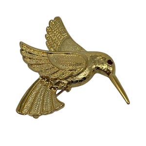 May include: A gold-toned hummingbird brooch with a red rhinestone eye. The hummingbird is in flight with its wings spread.