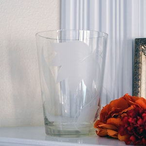 May include: A clear glass vase with an etched leaf design. The vase is sitting on a white surface with orange flowers in the foreground.