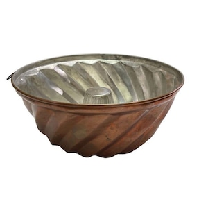 May include: A copper-colored bundt cake pan with a fluted design. The interior is silver-colored, and the pan has a central tube. The pan is designed for baking cakes and other desserts.