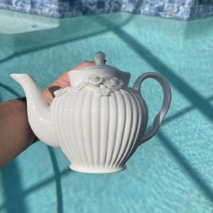 May include: A white ceramic teapot featuring a vertical ribbed texture on the body and a lid adorned with sculpted floral details. The teapot has a curved spout and a handle, with a smooth finish throughout the entire piece, showcasing a classic design.