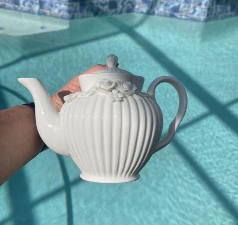 May include: A white ceramic teapot featuring a vertical ribbed texture on the body and a lid adorned with sculpted floral details. The teapot has a curved spout and a handle, with a smooth finish throughout the entire piece, showcasing a classic design.