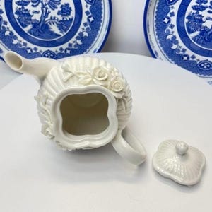 May include: A cream-coloured ceramic teapot with a fluted body and raised rose floral relief details. The teapot features a four-lobed opening, a curved spout, and a matching removable lid with a small knob handle, presented from a top-down view.