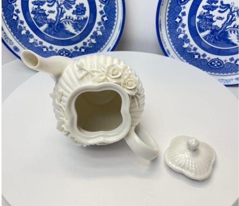May include: A cream-coloured ceramic teapot with a fluted body and raised rose floral relief details. The teapot features a four-lobed opening, a curved spout, and a matching removable lid with a small knob handle, presented from a top-down view.