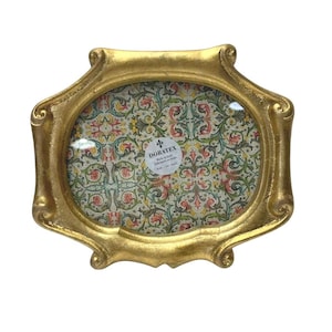 Peut inclure: Cadre photo ovale doré avec un design orné et texturé. L'intérieur présente un tissu à motifs avec des motifs floraux et de volutes dans les tons de vert, de rouge et de jaune. Le cadre porte le texte "DORATEX".