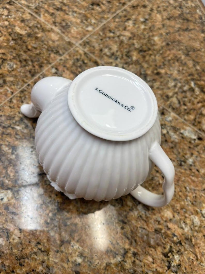 May include: A white ceramic teapot with a ribbed, vertical texture and a curved handle. The base of the teapot is visible, featuring the black printed text L.GODINGER & CO. along with a registered trademark symbol on the smooth circular bottom.