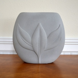 May include: A gray ceramic vase with a stylized flower design. The vase has a rounded shape and a smooth, matte finish.