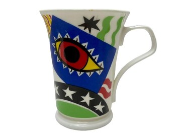 Puede incluir: Taza de cerámica blanca con un diseño abstracto colorido que presenta un ojo grande con acentos rojos, amarillos y negros. El diseño también incluye estrellas, rayas y líneas onduladas.