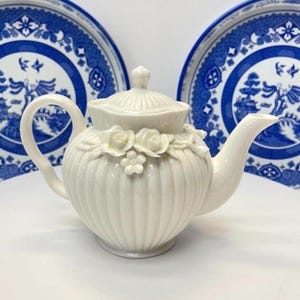 May include: A cream-coloured ceramic teapot featuring a vertical ribbed body, a matching lid, and a decorative relief of sculpted roses and small flowers around the upper section. The teapot has a curved handle and a tapered spout.