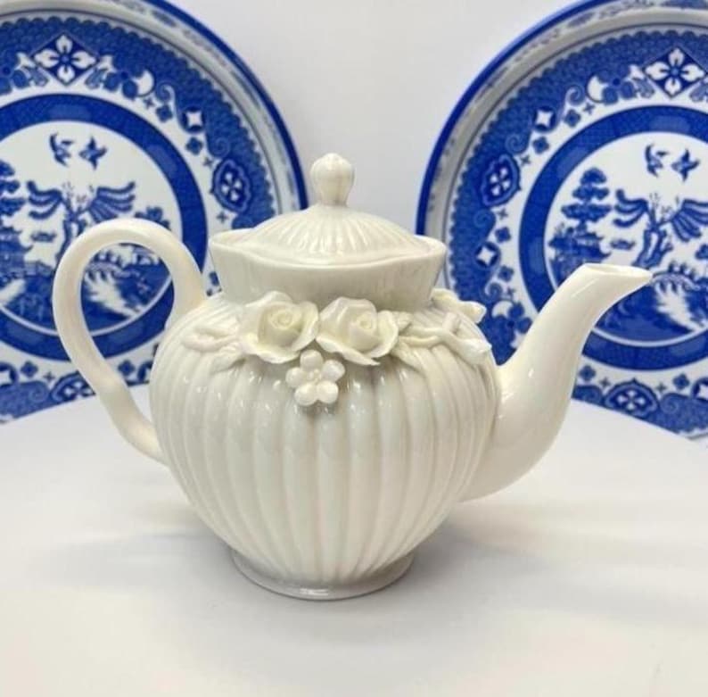 May include: A cream-coloured ceramic teapot featuring a vertical ribbed body, a matching lid, and a decorative relief of sculpted roses and small flowers around the upper section. The teapot has a curved handle and a tapered spout.