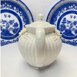May include: A cream-coloured ceramic teapot with a ribbed body, a matching lid, and sculpted floral accents on the sides. The teapot features a curved handle and a spout, presented in front of two blue and white patterned ceramic plates.