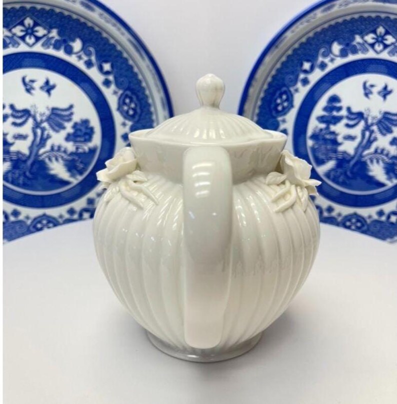 May include: A cream-coloured ceramic teapot with a ribbed body, a matching lid, and sculpted floral accents on the sides. The teapot features a curved handle and a spout, presented in front of two blue and white patterned ceramic plates.