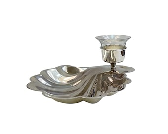 May include: A silver-toned metal tray shaped like a large scallop shell with a smooth, reflective surface. Attached to one side of the tray is a small, elevated silver-toned metal candle holder featuring a clear glass insert for a candle.