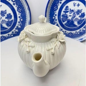 May include: A white ceramic teapot featuring a ribbed body and a matching lid. The teapot is decorated with sculpted white rose blossoms and leaves on the sides. Two blue and white patterned plates are positioned in the background.