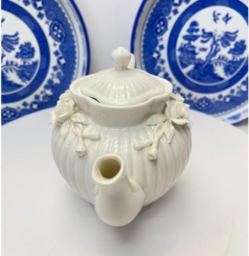 May include: A white ceramic teapot featuring a ribbed body and a matching lid. The teapot is decorated with sculpted white rose blossoms and leaves on the sides. Two blue and white patterned plates are positioned in the background.