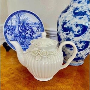May include: A white ceramic teapot with a ribbed body, a matching lid, and a decorative relief of three roses and small berries on the front. The teapot features a curved handle and a tapered spout, showcasing a glossy finish and elegant design.