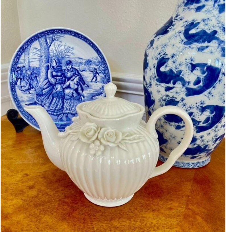 May include: A white ceramic teapot with a ribbed body, a matching lid, and a decorative relief of three roses and small berries on the front. The teapot features a curved handle and a tapered spout, showcasing a glossy finish and elegant design.