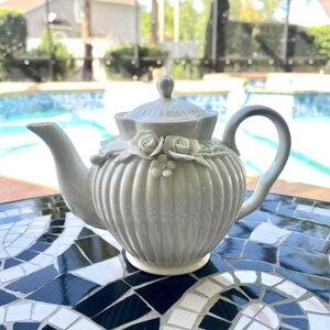May include: A light grey ceramic teapot featuring a ribbed, vertical texture and a lid with a small knob. The body of the teapot is adorned with sculpted, three-dimensional floral details, including two roses and a small flower centred on the front.