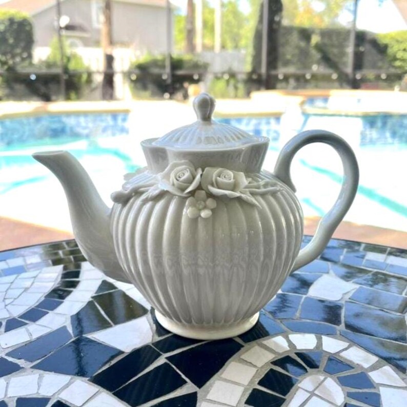 May include: A light grey ceramic teapot featuring a ribbed, vertical texture and a lid with a small knob. The body of the teapot is adorned with sculpted, three-dimensional floral details, including two roses and a small flower centred on the front.