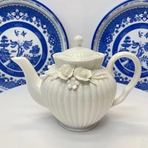 May include: A white ceramic teapot with a ribbed body and a lid, featuring three-dimensional sculpted rose and small flower decorations on the front. The teapot has a curved handle and a spout, positioned in front of two blue and white patterned plates.