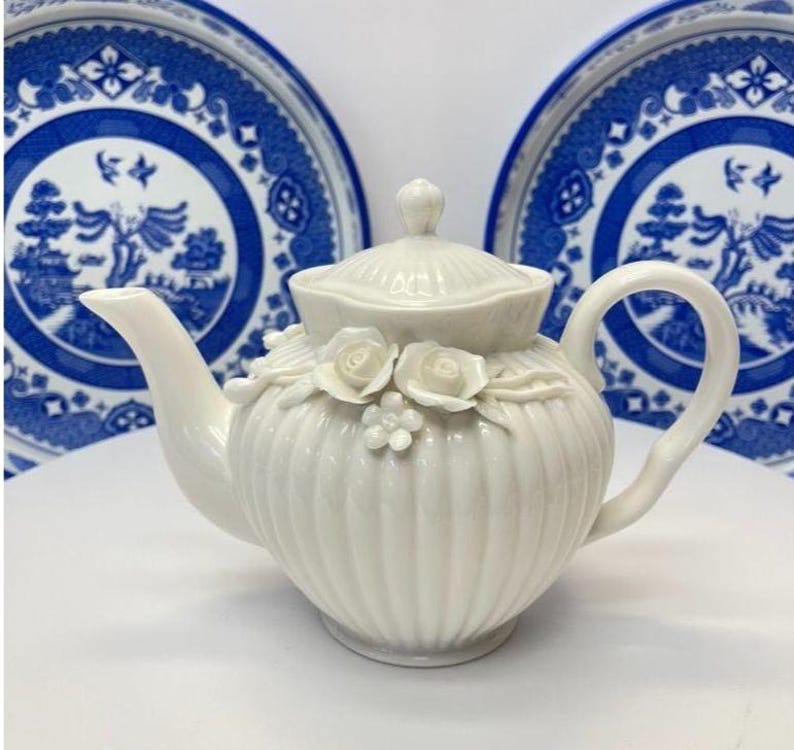 May include: A white ceramic teapot with a ribbed body and a lid, featuring three-dimensional sculpted rose and small flower decorations on the front. The teapot has a curved handle and a spout, positioned in front of two blue and white patterned plates.