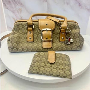 May include: A tan and brown patterned handbag with a central buckle, dual handles, and a matching wallet. The bag features a silver-toned charm with dice and a pearl-like bead attached to the handle, showcasing a geometric square print design.