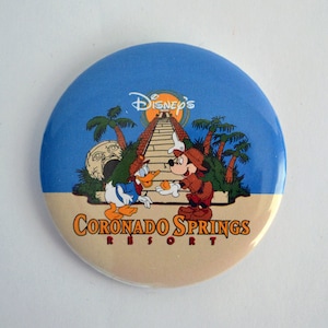 May include: A blue and beige round button featuring a cartoon image of Mickey Mouse and Donald Duck in front of a pyramid. The text "Disney's Coronado Springs Resort" is printed on the button.