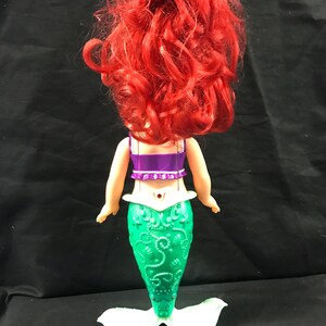 May include: A doll of a mermaid, viewed from the back. The doll has bright red hair, a purple top, and a green tail with a white fin. The tail has a detailed, textured design. The doll is standing on a white base.