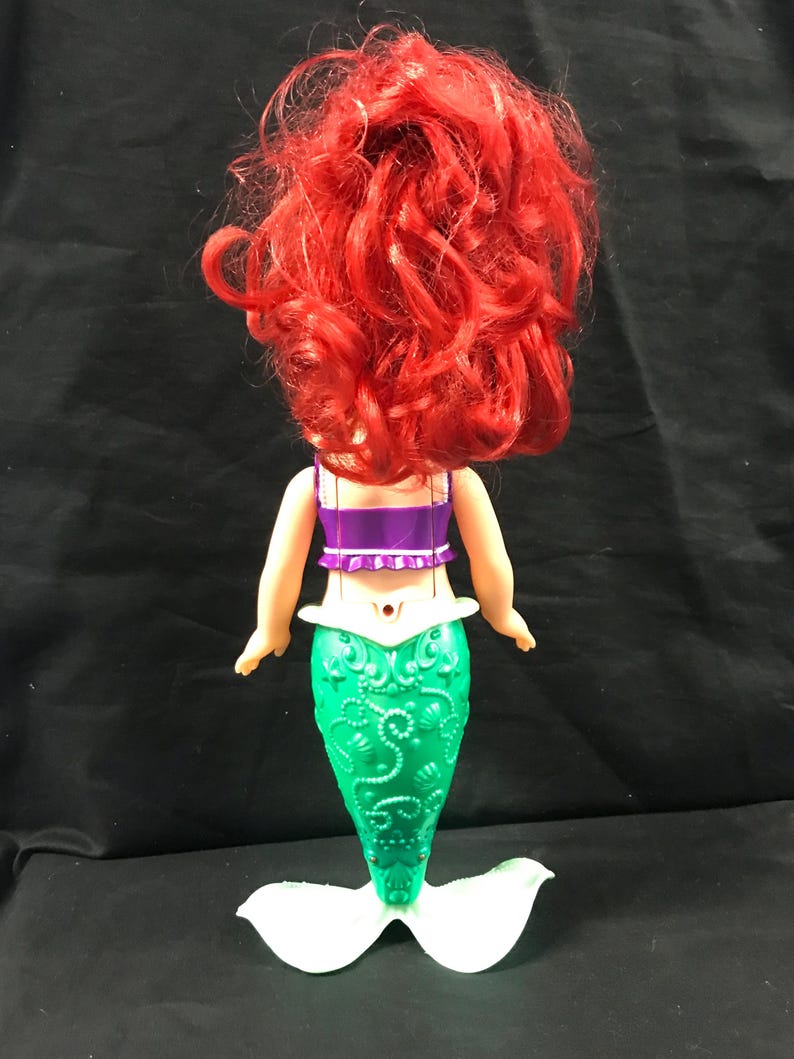 May include: A doll of a mermaid, viewed from the back. The doll has bright red hair, a purple top, and a green tail with a white fin. The tail has a detailed, textured design. The doll is standing on a white base.