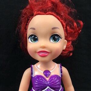 May include: A doll with red hair and large green eyes. The doll is wearing a purple seashell top and a white necklace with a seashell pendant. The doll has a light skin tone and a pink lipstick.