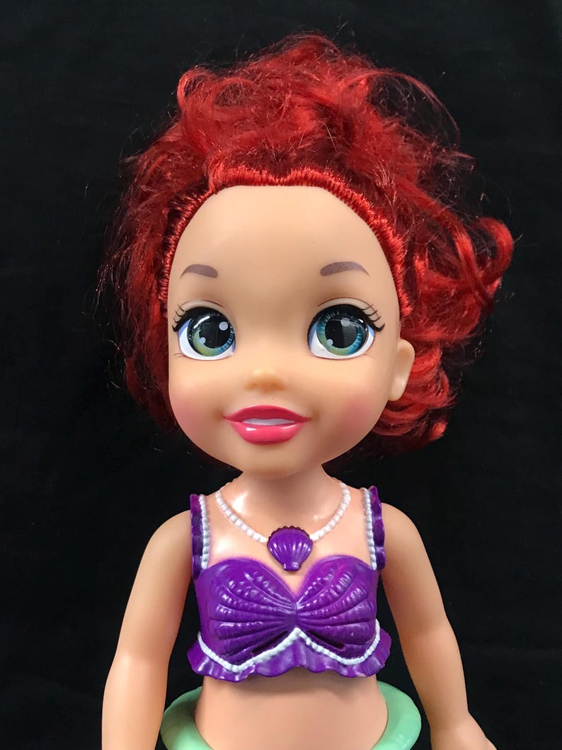 May include: A doll with red hair and large green eyes. The doll is wearing a purple seashell top and a white necklace with a seashell pendant. The doll has a light skin tone and a pink lipstick.